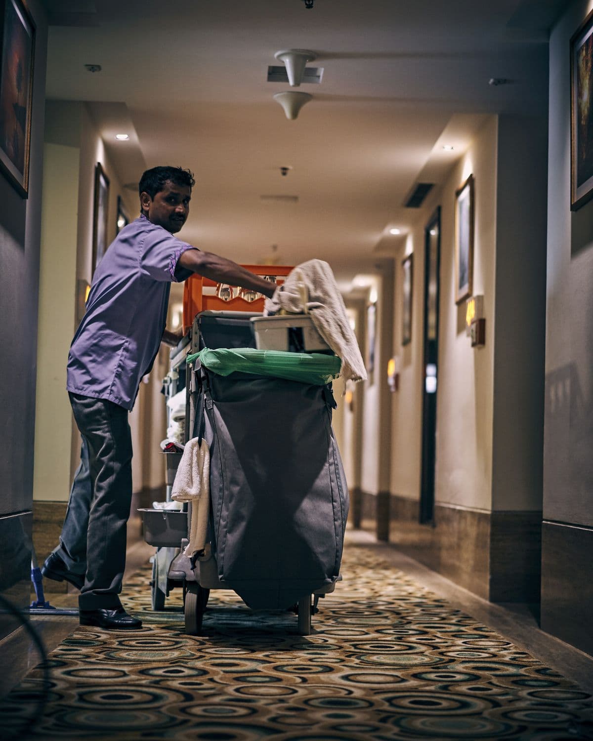 Een housekeepingmedewerker in uniform duwt een schoonmaakkar door een hotelgang.