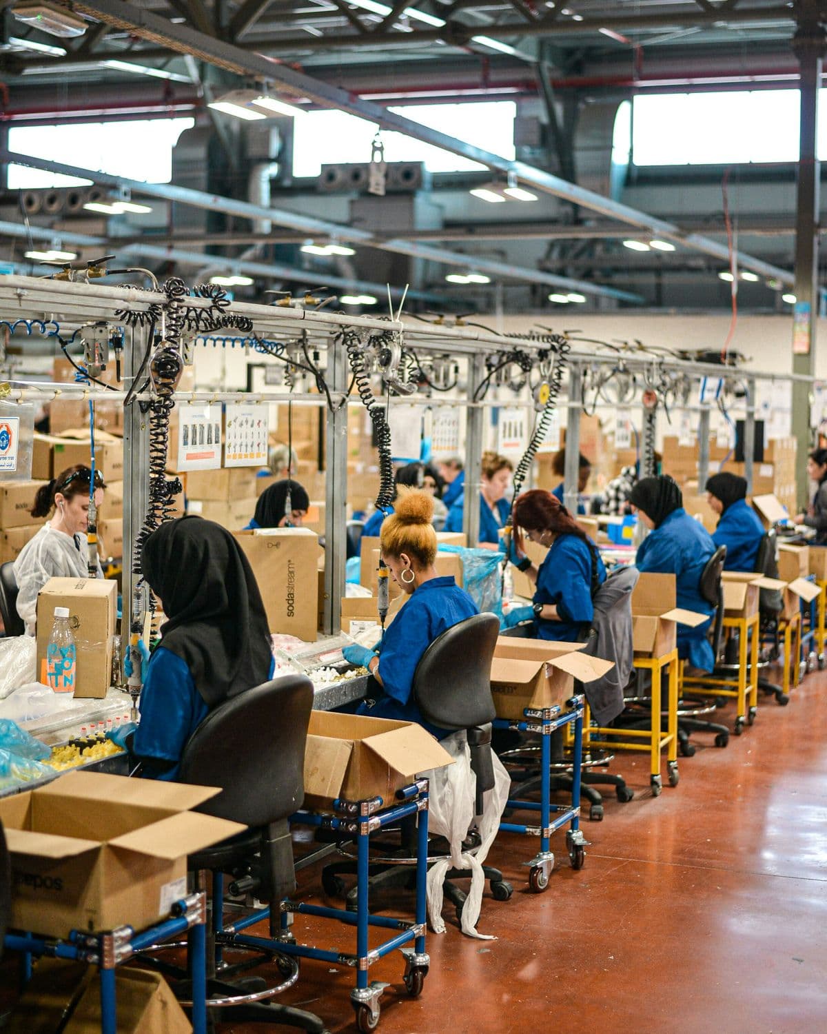 Medewerkers in blauwe werkkleding aan een lange werkbank in een fabriekshal, tussen kartonnen dozen.