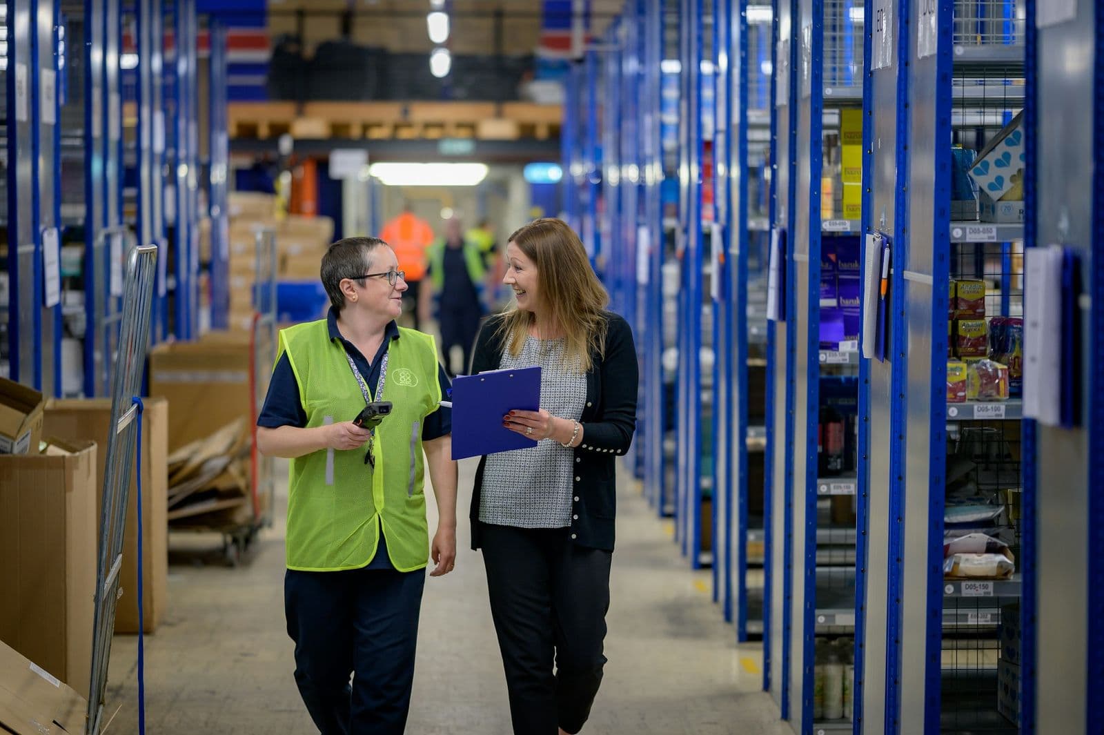 Een medewerker in een geel hesje met een handscanner loopt in gesprek met een collega door een magazijngang.