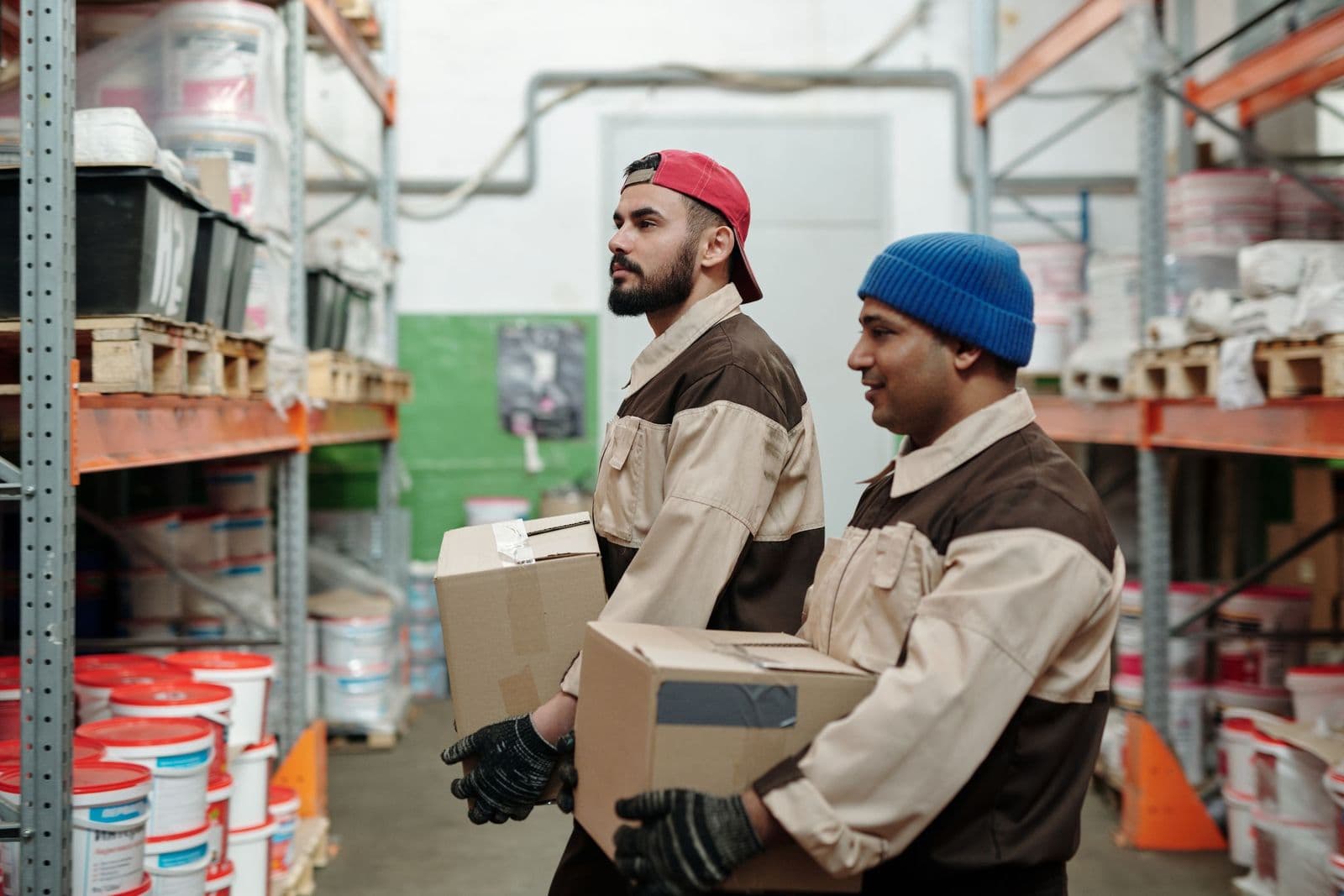 Twee magazijnmedewerkers in werkjassen dragen samen kartonnen dozen tussen de stellingen.