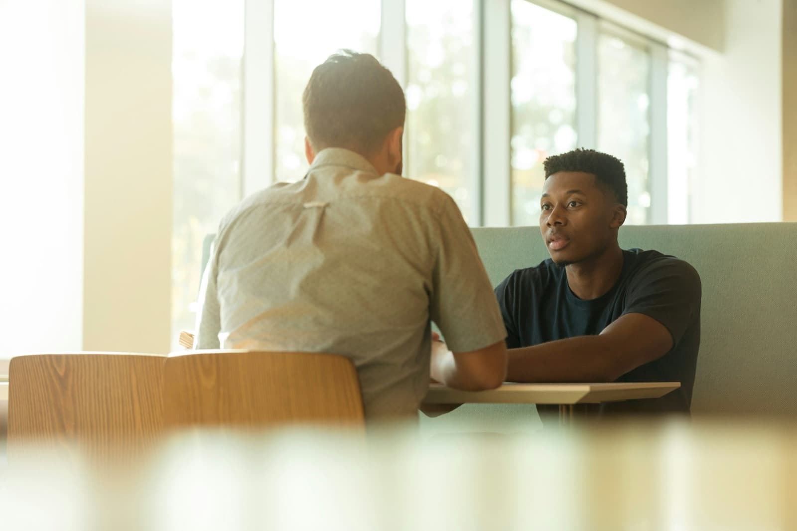 Twee mannen in gesprek aan een tafel bij een raam, in zacht tegenlicht.